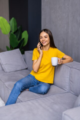 Lovely young woman sitting on a floor at home, drinking tea, leaning on a couch, talking on mobile phone