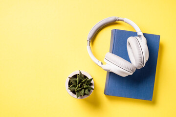 Audio book concept. Headphones, book and green plant at yellow background. Flat lay image with copy space.