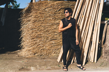 Young man standing near hay and bamboo stack in sun