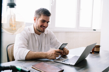 Smiling man chatting on smartphone during work via laptop