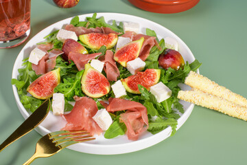 Autumn figs salad with arugula, feta, raw ham in white plate on light green background served with rose wine and grissini. Flexitarian diet, paleo diet fall or summer salad, close up,