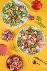 Autumn figs salad with arugula, feta, raw ham in two white plate on yellow background served with rose wine. Flexitarian diet, paleo diet fall or summer salad, top view, flat lay, vertical