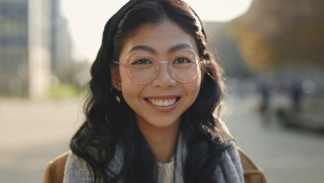 Portrait of the Attractive Smiled Asian Woman Wearing Glasses Looking at Camera Standing Outside. People Face. Girl Looking at the Camera on the City Background. Young Business Woman Outside