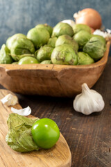 Tomatillo with papery husk removed on a cutting board. Wooden bowl with Tomatillos, garlic, and onions in background. 