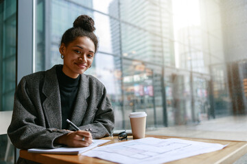 Woman interior designer making notes while sitting in cozy cafe near window and looking at camera