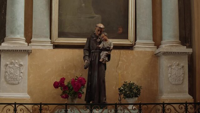 Statue of Saint Anthony with child in his arms stands in old church near wall with cracks. The columns are decorated with stucco moldings with coat of arms.