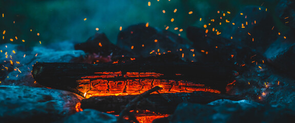 Vivid smoldered firewoods burned in fire close-up. Atmospheric background with orange flame of campfire and blue smoke. Warm full frame image of bonfire. Glowing embers in air. Bright sparks in bokeh