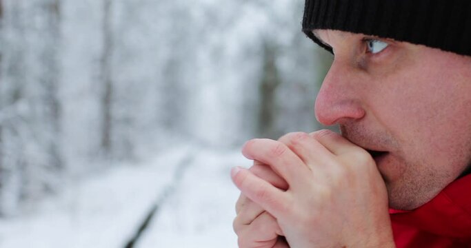 Man frozen in the winter forest. Close up male warms his hands breathes with steam from his mouth on them and rubs on frosty winter day, in the background snowy trees. Holiday vacation trip.