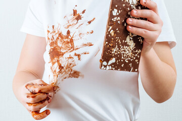 A child's hand showing a dirty t-shirt with a chocolate stain and holding a bar of chocolate in the...
