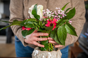 Close-up woman with jeans and beige pullover is putting bouquet of flowers in a vase indoors