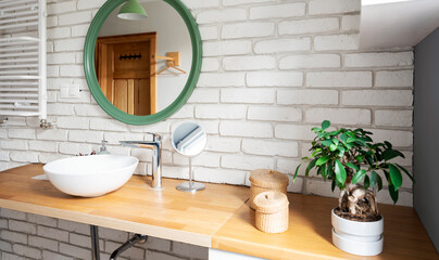 Bathroom interior with round mirror on brick wall and ceramic washbasin on wooden counter. 