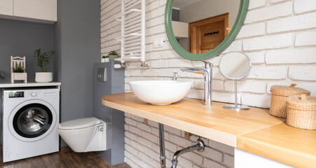 Modern bathroom interior with ceramic wash basin on wooden counter with faucet and round mirror on white brick wall. Banner.