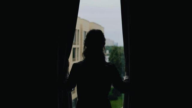 The girl stands in front of the window and pushes the curtains in front of her. There is a view of the street