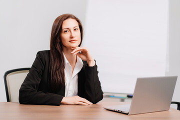 brunette woman in black coat in office with meeting board, desktop and laptop 