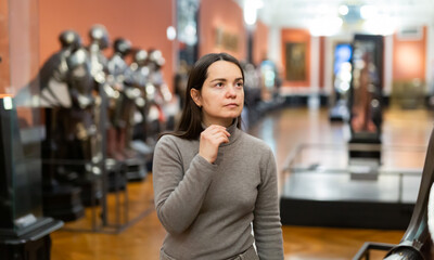 Female museum visitor examining with interest ancient iron armor displayed on exhibition ..
