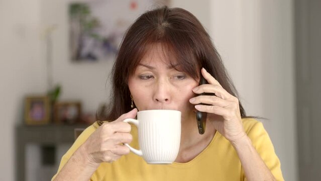 Frontal closeup beautiful mature woman long hair thin earrings in yellow top talking on mobile phone smiling drinking hot coffee winces face. 