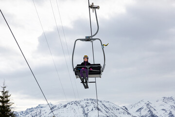 Smiling woman skier in full ski equipment riding on chairlift, lifting to top of mountain to pistes on background of snowy peaks on cloudy winter day..