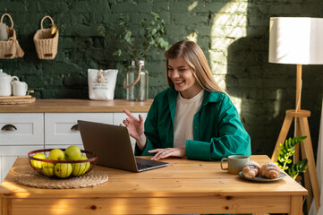 A young smiling woman is chatting with someone online via video call on a laptop while sitting at a wooden table in the kitchen.