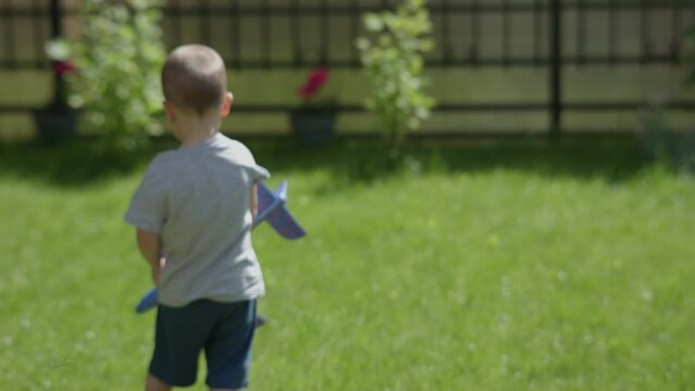 little boy child playing with airplane garden sunny summer day. male kid boy grey t-shirt pretending pilot flying blue toy plane green grass outdoors with bright flowers background. outdoor activity
