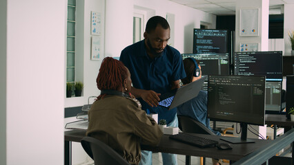 Obraz premium Two software coders analyzing source code looking for errors on laptop, checking big tv monitor in it agency office. Programmers working on cyber security innovation in busy company space.