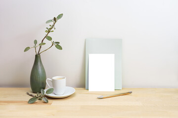 Blank greeting card or invitation mock-up with a pen next to a gray green vase with sage branches and a coffee cup, simple home decor style, copy space