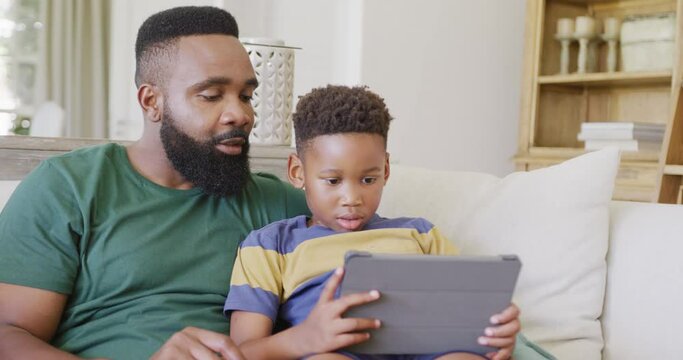 Happy african american father and son sitting on sofa, using tablet, in slow motion