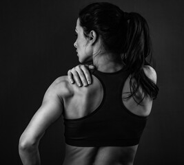 Fototapeta na wymiar Female sporty muscular with ponytail doing stretching workout of the shoulders, blades in sport bra, holing the neck the hand on dark grey background with empty copy space. Back view. Banner