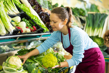 Positive middle-aged saleswoman putting fresh cauliflowers on food stall in grocery store