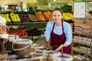 Smiling middle-aged shop assistant politely showing olives in ladle in big greengrocery