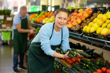 Smiling middle-aged shop assistant laying fresh cucumbers on counter in big greengrocery
