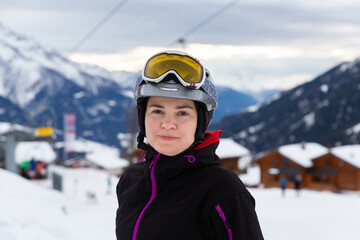 Fototapeta na wymiar Young woman spending winter holidays in ski resort, posing in ski helmet and goggles outdoors with snow covered mountains in background