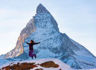 Young female traveler enjoying winter holidays in Swiss Alps, jumping with raised hands in nature with snowy sharp Matterhorn peak in background