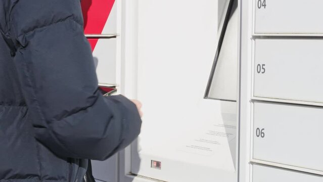 A young caucasian man in a blue jacket enters a code with his finger on the screen of a mailbox to take a parcel, close-up side view. The concept of receiving a parcel, a modern lifestyle.
