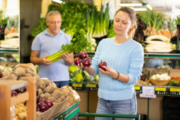 Positive middle-aged woman purchaser taking red onions at the counter in grocery store