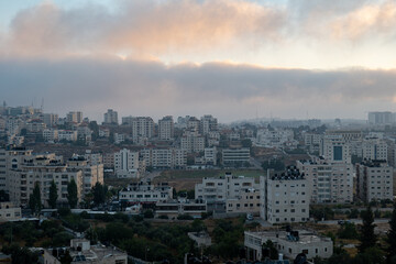 Fototapeta na wymiar Dark Ramallah Cityscape at Dawn with High White Buildings and Trees Against Cloudy Sky