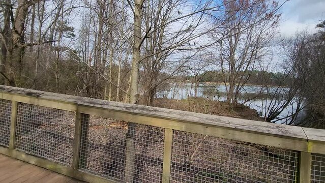panning footage along a brown wooden bridge with bare winter trees, green trees and a lake with blue sky and clouds at Rhodes Jordan Park in Lawrenceville Georgia USA