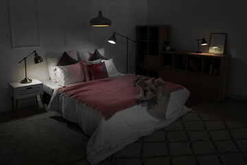 Interior of bedroom with blankets on bed and glowing lamps at night