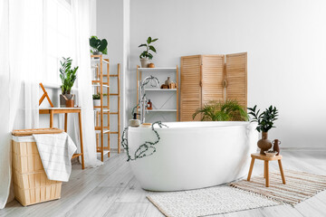 Interior of light bathroom with bathtub, shelving units and houseplants