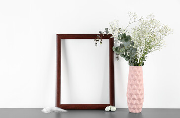 Blank frame, vase with flowers, Easter eggs and feather on table near white wall
