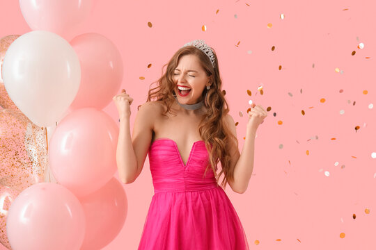 Happy young woman in prom dress with balloons and confetti on pink background