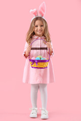 Cute little girl in bunny ears with basket of Easter eggs on pink background
