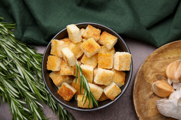 Delicious crispy croutons in bowl and rosemary on dark table, flat lay