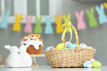 Easter basket with tasty painted eggs near cake and figure of rabbits on white table indoors