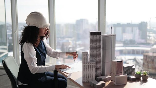 Curly female engineer working on building complex prototype project of residential or business district, measuring blueprint city model using compass at office with panoramic windows