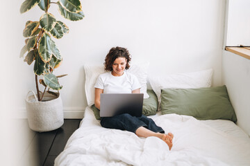 Young brunette woman in home clothes working on laptop on bed