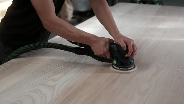 young man builder carpenter grinds a wooden table with a grinding machine in the shop. A construction worker using a cordless power sander to sand a massive wooden table top