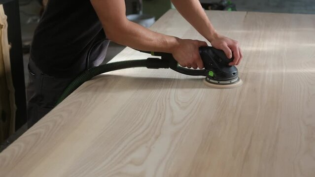 young man builder carpenter grinds a wooden table with a grinding machine in the shop. A construction worker using a cordless power sander to sand a massive wooden table top