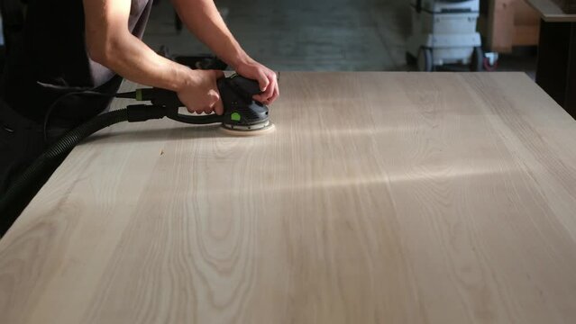 young man builder carpenter grinds a wooden table with a grinding machine in the shop. A construction worker using a cordless power sander to sand a massive wooden table top
