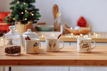 White ceramic mugs and a teapot stand on a wooden table against the backdrop of a kitchen, glowing garlands and a Christmas tree. Family breakfast at home.