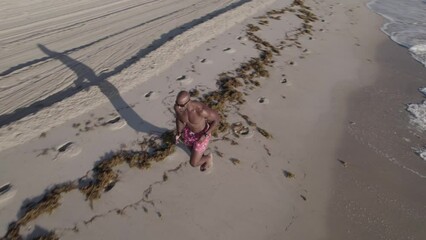 Drone orbiting footage of determinate muscular male athlete running down a shoreline on a sunny day